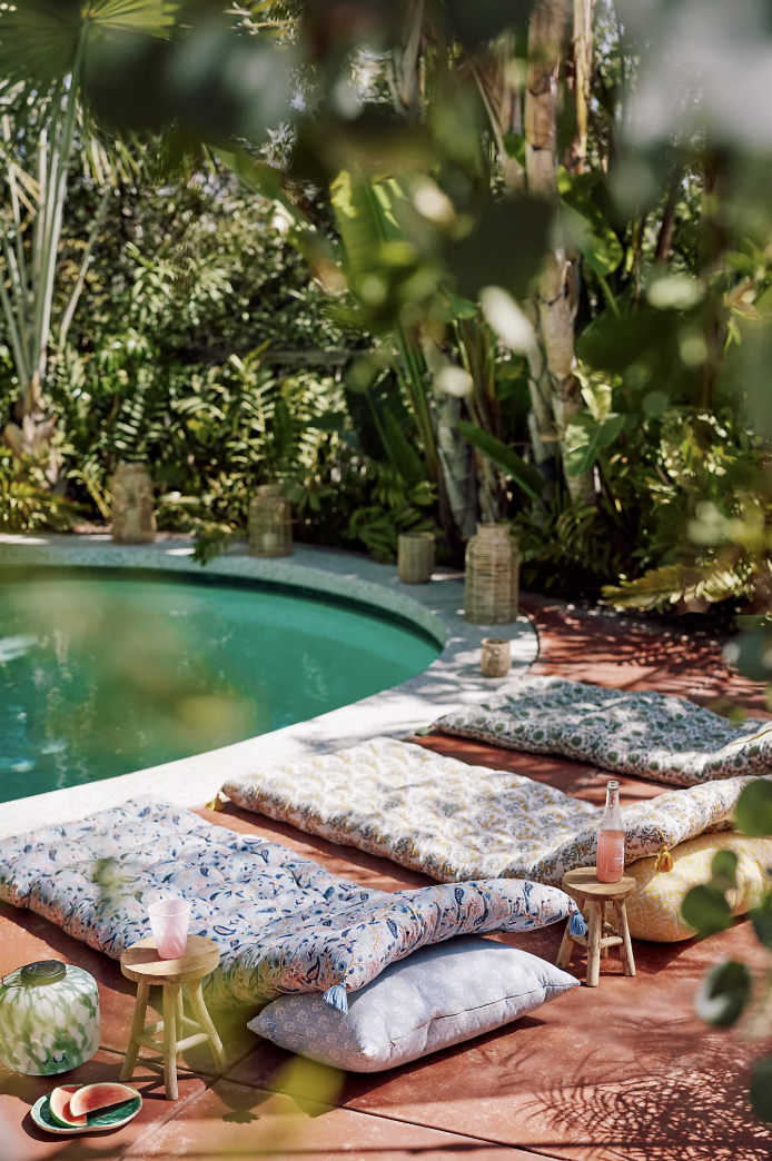 A lush tropical backyard scene featuring a round pool surrounded by leafy greenery. Colorful patterned floor cushions are laid out on a terracotta patio, paired with small wooden stools holding pink drinks and ceramic dishes. Rattan lanterns are scattered along the pool’s edge, creating a cozy, boho-inspired outdoor lounge setup.